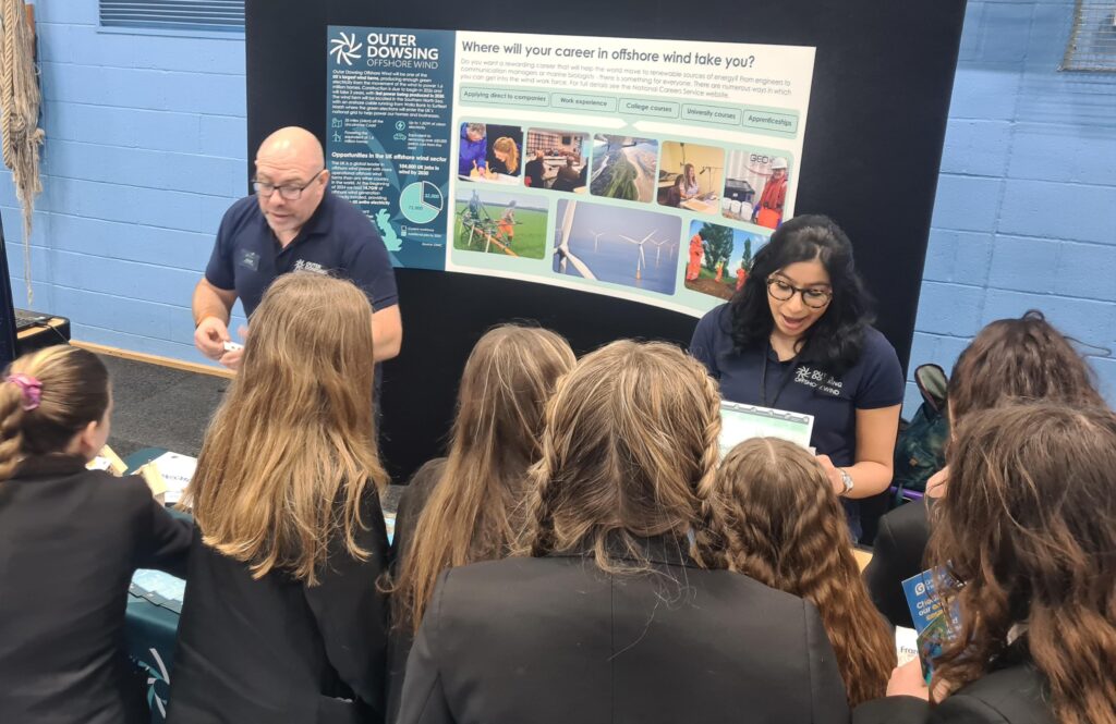 Two Outer Dowsing Offshore Wind Engineers in blue polo shirts behind an exhibition table talking to 10 girls about the wind farm. 