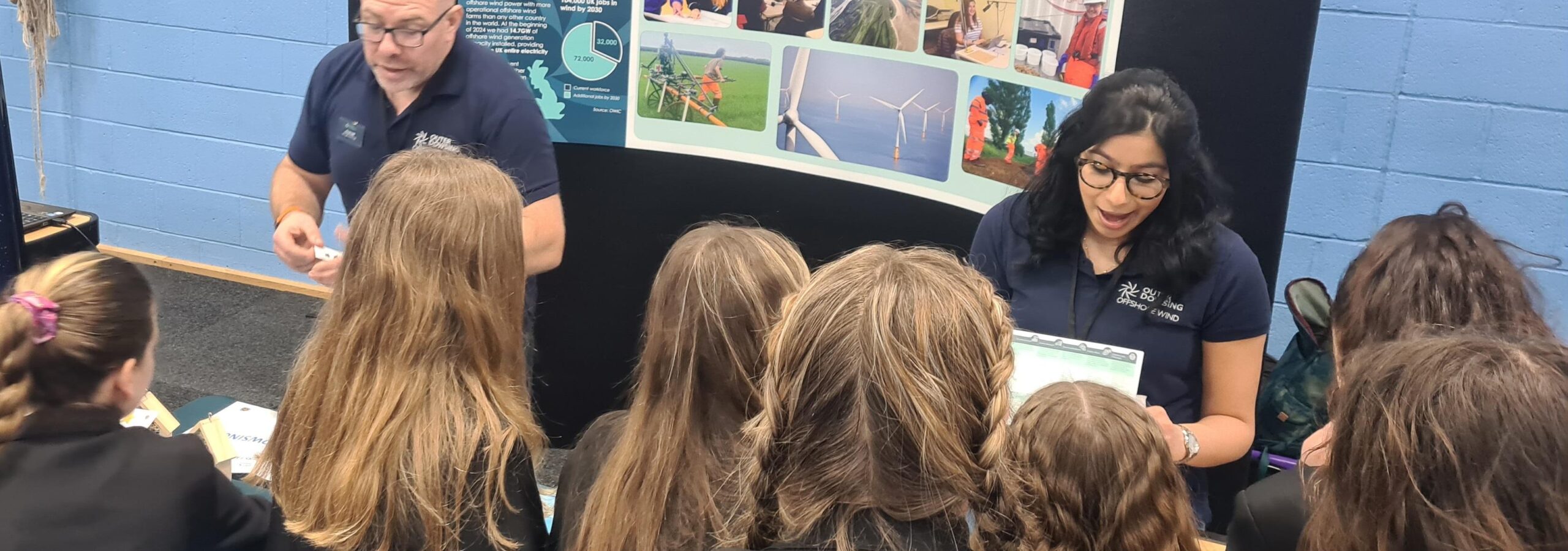 Two Outer Dowsing Offshore Wind Engineers in blue polo shirts behind an exhibition table talking to 10 girls about the wind farm.