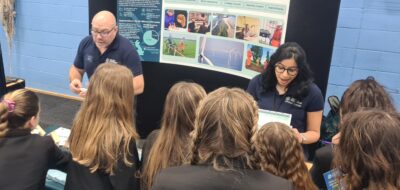 Two Outer Dowsing Offshore Wind Engineers in blue polo shirts behind an exhibition table talking to 10 girls about the wind farm.