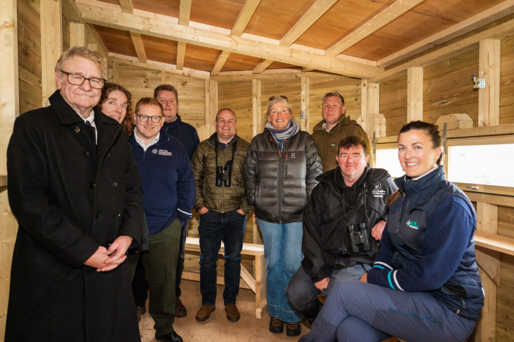 A group of people who attended the bird hide opening inside the bird hut wearing jackets, jeans and a few with binoculars at the opening of the bird hide.